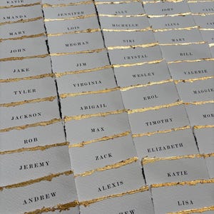 May include: A collection of place cards with gold foil accents and names written in black ink. The cards are arranged in rows and columns on a gray surface.