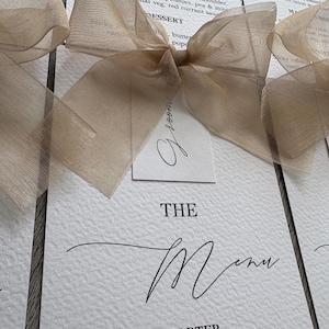 Może przedstawiać: Zbliżenie kart menu weselnego z beżową kokardą. Na kartach znajdują się słowa "THE MENU" i "STARTER" wydrukowane czarnym skryptem. Menu zawiera dania takie jak parfait z wątróbki drobiowej i łopatka jagnięca.