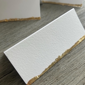 Peut inclure: Cartes de table blanches avec un bord en feuille d'or. Les cartes sont rectangulaires et ont une surface texturée. Plusieurs cartes sont visibles, la feuille d'or ajoutant une touche d'élégance.