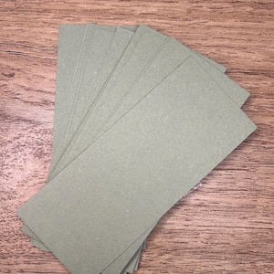May include: A collection of rectangular, sage green paper cards fanned out on a wooden surface. The cards are evenly cut and have a slightly textured appearance. The wood grain background provides a natural contrast to the cards.