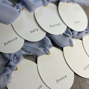 Op de afbeelding: Close-up van verschillende witte ovale naambordjes met namen in het zwart gedrukt, bevestigd aan lichtblauwe zijden linten. Elke kaart heeft een gouden oogje. Namen zijn onder meer Rebecca, Sally, Hannah, John, Andrew en Jessica. De kaarten zijn gerangschikt op een houten oppervlak.