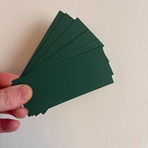 Puede incluir: Una mano sostiene un abanico de tarjetas rectangulares de color verde oscuro. Las tarjetas son de un color verde oscuro sólido y parecen estar hechas de papel texturizado. El fondo es una pared lisa de color blanco roto.