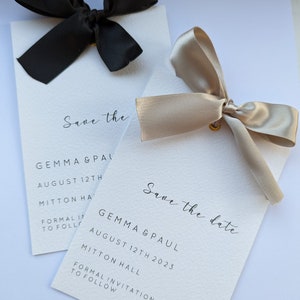 May include: Two save the date cards with black and beige bows. The text on the cards reads "Save the date" and includes the names Gemma & Paul, the date August 12th, 2023, and the location Mitton Hall. Formal invitation to follow.