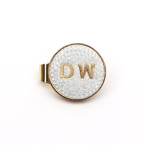 May include: A gold-toned metal tie clip with a circular face. The face has a textured, opalescent background and the gold letters "DW". The clip is designed to secure a tie to a shirt.
