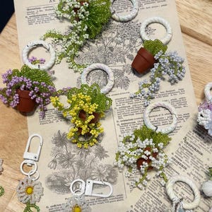 May include: A collection of handmade beaded keychains and charms featuring miniature potted plants and daisy designs. The keychains have white metal clasps and braided white rings. The background includes pages from a vintage botanical dictionary.