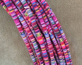 May include: Close-up of several colorful, woven cords. The cords are primarily pink and purple, with accents of blue, green, yellow, and white. The cords have a textured appearance and are arranged in a bundle. The background is a neutral, tan color.