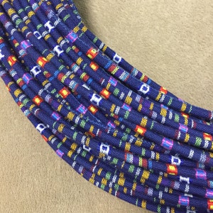 May include: Close-up of a bundle of navy blue fabric cords with a colorful plaid pattern. The pattern includes squares of red, yellow, green, and white. The cords are arranged in a curved shape against a tan background.