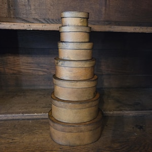 May include: A stack of six round, light brown wooden boxes with lids, arranged in descending order of size from the bottom to the top. Each box features a smooth, cylindrical shape with a visible grain and a consistent, natural wood finish.