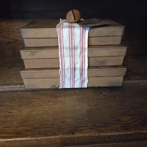 May include: A stack of three rectangular brown metal boxes with a distressed finish. A strip of white fabric with thin red vertical stripes is wrapped around the center of the stack. A small, round, rusted metal bell sits on top of the boxes.