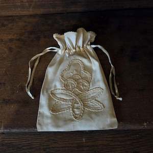 May include: A cream-colored fabric drawstring pouch featuring a decorative embroidered floral motif on the front. The bag is cinched at the top with matching fabric ribbons, creating a gathered edge. The embroidery consists of stitched outlines.