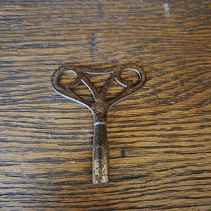 May include: A vintage metal clock winding key featuring a decorative, open-work butterfly-shaped handle with a weathered, dark patina. The stem is cylindrical and tapers slightly toward the base, showing signs of age and oxidation on the surface.