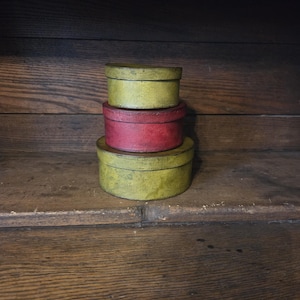 May include: A stack of three round, lidded boxes with a distressed finish. The top and bottom boxes are a muted olive green, while the middle box is a deep, weathered red. Each container features a simple cylindrical shape with a flat lid.