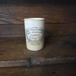 May include: A vintage cylindrical marmalade jar with a cream-colored, cracked glaze. The jar features a black and white label with the text "JAMES KEILLER & SONS DUNDEE MARMALADE" and "PRIZE MEDAL FOR MARMALADE LONDON, 1862".