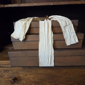 May include: Three stacked brown rectangular boxes with a cream-colored fabric draped over them. The boxes are on a wooden surface, and the background is a dark wooden cabinet.