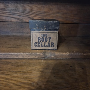 May include: A small, rectangular box with a dark, distressed finish. The box features a label with the text "1863 ROOT CELLAR" in a vintage font. The box is set against a wooden background.