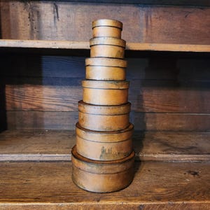 Puede incluir: Una pila de siete cajas antiguas redondas con tapa de diferentes tamaños. Las cajas están hechas de un material marrón claro con bordes más oscuros y están dispuestas sobre una superficie de madera. Las cajas están apiladas unas sobre otras.