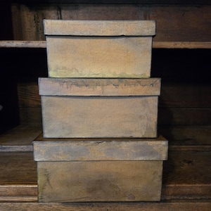 May include: Three stacked, rectangular boxes with lids. The boxes are a muted, earthy brown with a slightly distressed appearance. They are arranged on a wooden surface, suggesting storage or display.