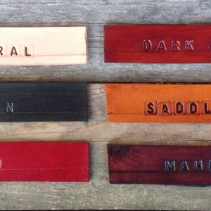 Pu&ograve; includere: Sei campioni di pelle in diversi colori e finiture. I campioni sono etichettati "Naturale", "Marrone scuro", "Verde", "Sella Tan", "Rosso" e "Marrone".