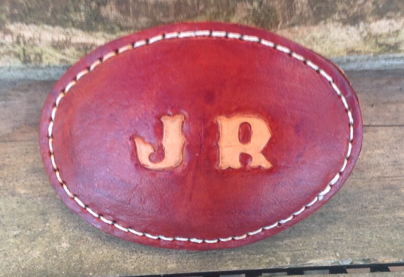 Pu&ograve; includere: Una fibbia per cintura in pelle rossa con cuciture bianche e le iniziali "JR" in rilievo in color marrone chiaro.