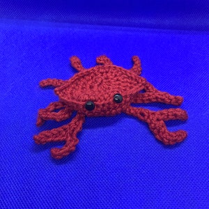 May include: A handmade crochet crab in a deep red color, featuring black safety eyes. The crab has multiple legs and a textured body, set against a solid blue background. This craft item is a unique decorative piece.