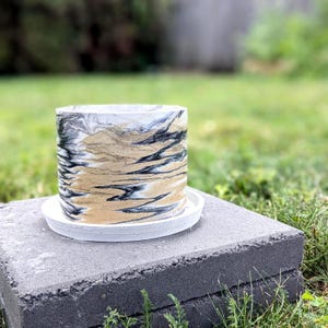 May include: A cylindrical planter featuring a marbled pattern of black, white, and metallic gold swirls. The planter sits on a matching circular white saucer, creating a cohesive set for holding plants with a smooth, decorative exterior finish.