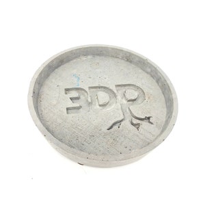 May include: A round, gray concrete coaster with the word "EDR" embossed in the center. The "R" is stylized as a tree with roots.