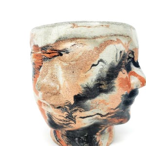 May include: A ceramic planter shaped like a face with abstract black, white, and orange marbling. The planter has a wide opening at the top and a rounded bottom.