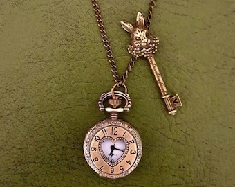 May include: A gold-tone necklace with a pocket watch and a key charm. The pocket watch has a heart-shaped face and Roman numeral markings. The key charm features a rabbit head.
