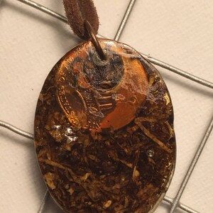 May include: A brown leather cord necklace with a pendant made from a copper coin encased in clear resin. The coin is visible through the resin and has the words "United States of America" and "One Cent" inscribed on it.