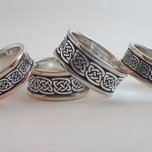 May include: Four silver rings with a Celtic knot design. The rings have a band of silver with a black background and a gold or rose gold inner band. The rings are displayed on a white surface.