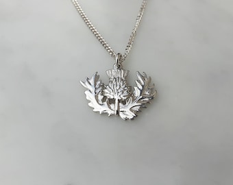 Puede incluir: Collar con colgante plateado que representa un cardo, símbolo de Escocia. El colgante, con hojas detalladas y una flor central, cuelga de una fina cadena de plata.