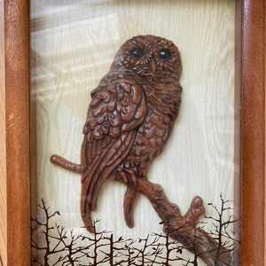 May include: A wooden owl sculpture perched on a branch, framed in a wood frame with a glass front. The owl is brown with black eyes and is set against a background of a forest silhouette.