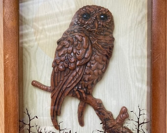 May include: A wooden owl sculpture perched on a branch, framed in a wood frame with a glass front. The owl is brown with black eyes and is set against a background of a forest silhouette.
