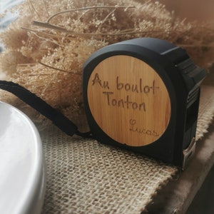 Può includere: Un metro a nastro nero e marrone con un viso in legno che dice "Au boulot Tonton Lucas".