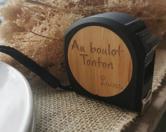 Peut inclure: Un mètre ruban noir et marron avec un visage en bois qui dit "Au boulot Tonton Lucas".
