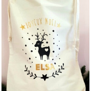 May include: A white canvas Christmas gift bag with a gold drawstring. The bag features a black reindeer with a heart, gold stars, and the text "Joyeux Noel" and "Elsa".