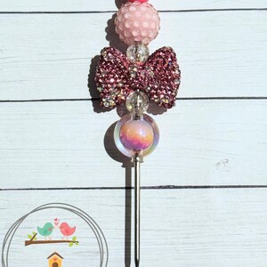 May include: A decorative cocktail pick with a silver top, pink beads, a sparkly bow, and an iridescent orb. The pick has two prongs and is designed for use in cocktails or appetizers. The image also includes a logo with birds and the text "2311 Decor & More."