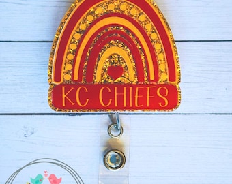 May include: Red, yellow, and gold glitter rainbow with a red heart in the center. The rainbow is on a red background with the text "KC CHIEFS" in white letters. The rainbow is attached to a clear retractable badge holder.