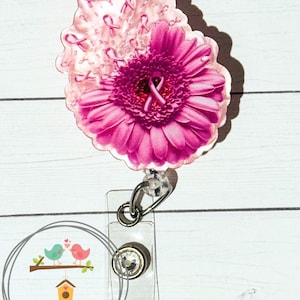 May include: A pink gerbera daisy badge reel with a pink ribbon design. The badge reel has a clear plastic holder and a metal clip. The text "2311 Decor & More" is in a circle at the bottom.