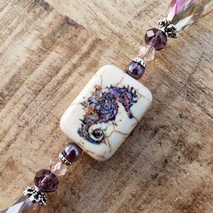 May include: A white rectangular charm with a purple seahorse design, attached to a beaded chain with purple and clear glass beads.