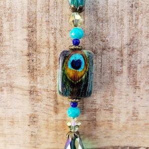 Może przedstawiać: Zawieszka w kształcie pióra pawia z wzorem w kolorze niebieskim, zielonym i złotym. Zawieszka jest nawleczona na naszyjnik z koralików w kolorze turkusowym, złotym i zielonym.