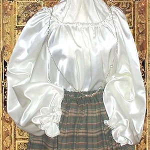 Pode incluir: Uma blusa branca de cetim com mangas compridas e bufantes e um decote franzido. A blusa &eacute; usada sobre uma saia xadrez.