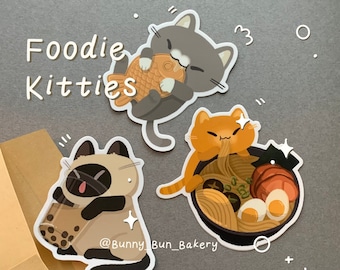 May include: Three colorful stickers featuring cartoon cats with food. One gray cat holds a fish-shaped pastry, an orange cat eats ramen, and a Siamese cat drinks boba tea. The text "Foodie Kitties" is at the top.
