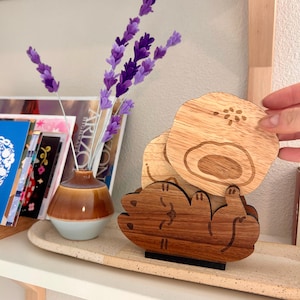 May include: Wooden coaster set featuring a bear design. The set includes a holder and two coasters with a light wood finish. The holder is a darker wood. A small vase with purple flowers is in the background.