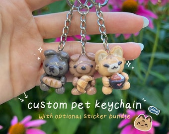 May include: Three custom pet keychain charms made of polymer clay. The charms are shaped like a grey bear holding a croissant, a brown bear holding a croissant, and an orange cat holding a bowl of ramen. The charms are attached to silver keychains.