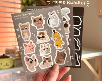 May include: A set of three sticker sheets featuring various cat memes. The first sheet is titled "Meme Cats Vol. 1" and features a variety of cats in different poses and expressions. The second sheet features a cat wearing a banana costume and a cat with a confused expression. The third sheet features a cat with a loading sign and a cat with a grumpy expression.