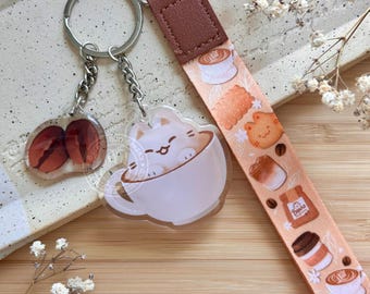 May include: Keychains and a lanyard with a coffee theme. One keychain features coffee beans, the other a cat in a coffee cup. The lanyard has a brown strap and a peach-colored ribbon with coffee and pastry illustrations. The text "Bunny Bun Bakery" is visible.