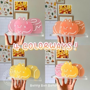Puede incluir: Cuatro conjuntos de adornos con forma de gato y donut en rosa, naranja, amarillo y rosa-naranja. Cada conjunto se muestra en una estantería transparente. El texto "4 COLORWAYS!" está en el centro. El logotipo de Bunny Bun Bakery está abajo a la izquierda.