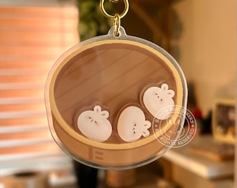 May include: A clear acrylic keychain featuring a cartoon illustration of three white bunny-shaped buns in a brown bamboo steamer. The text "Bunny Bun Bakery" is printed on the keychain.