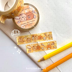 May include: A roll of yellow washi tape with a cartoon design of a bakery with the text "Crumbs and Maple Washi Tape" and "Bunny Bun Bakery".